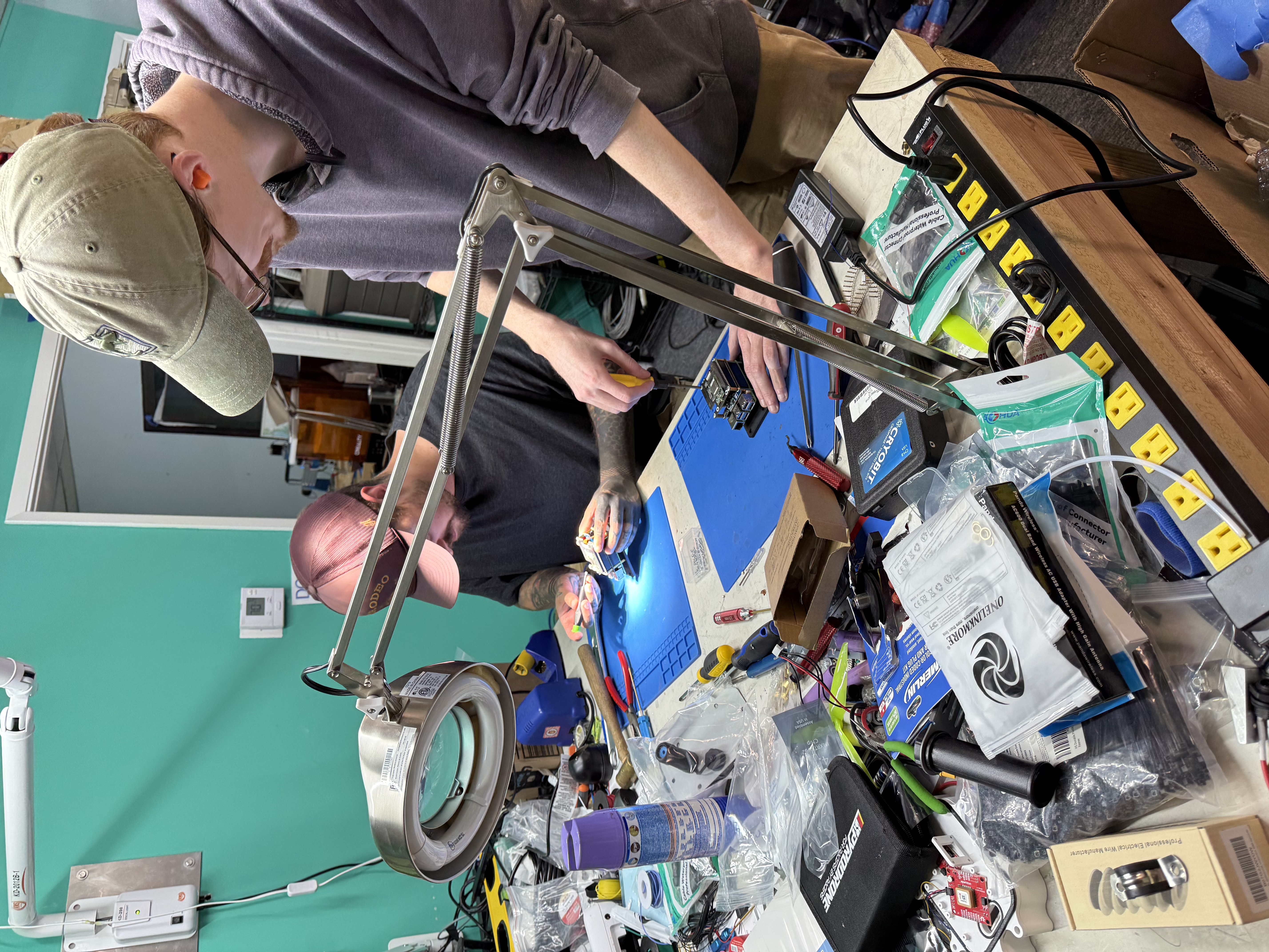 Two Iron Hornet engineers under magnifier lamps at the build bench, boards and tools across the workspace.